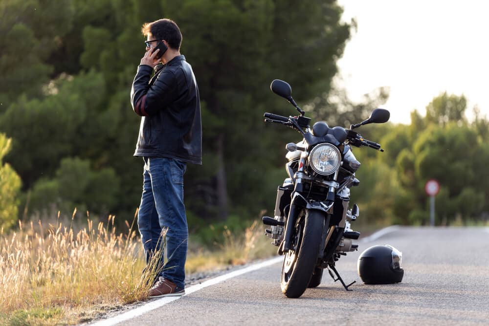 A man motorcyclist seeking affordable motorcycle insurance in CA contacts his insurer while dealing with a roadside breakdown.
