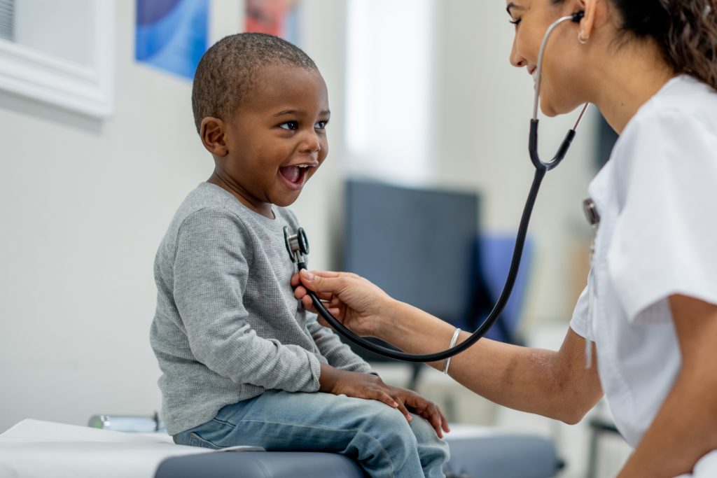 A young boy of African decent, sits up on an exam table as a female Pediatrician preforms a check-up on him.  The boy is dressed casually and smiling as the doctor listens to his heart.