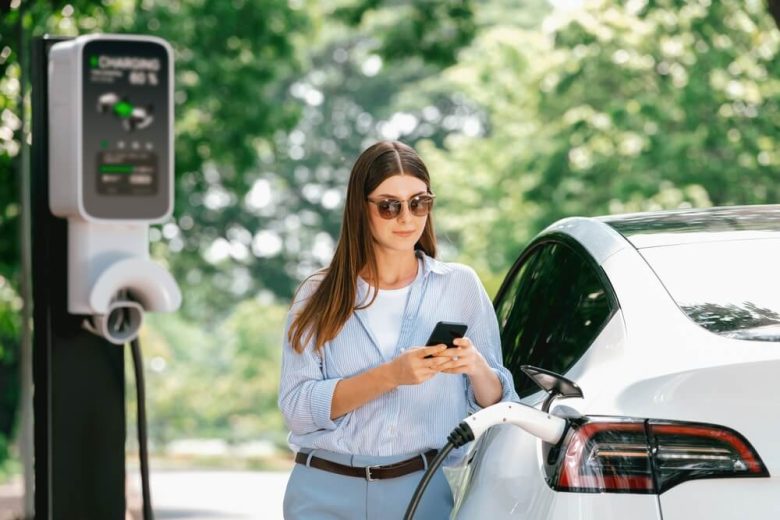 The young woman is using a smartphone online banking application to pay for charging the electric car battery at an EV charging station during her vacation holiday road trip, while also evaluating if hybrids are worth it.