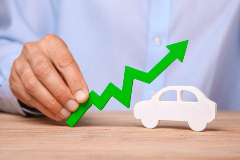 A picture illustrating rising car insurance prices with a man's hand holding an arrow going up over a small while car - cheap car insurance in California.